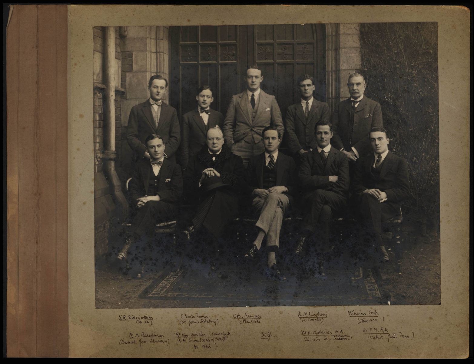 Photographer unknown. Group portrait, including Beverley Nichols, S. R. Sidebottom, J. Victor Evans, C. B. Ramage, R. M. Lindsay, William Gib, N. A. Beechman, Rt. Hon. Winston S. Churchill, W. H. [Noberly] M. A., D. P. M. Fyfe, circa 1920-1930.  Process: Smooth, matte gelatin silver print, on a gray-green paper board album page